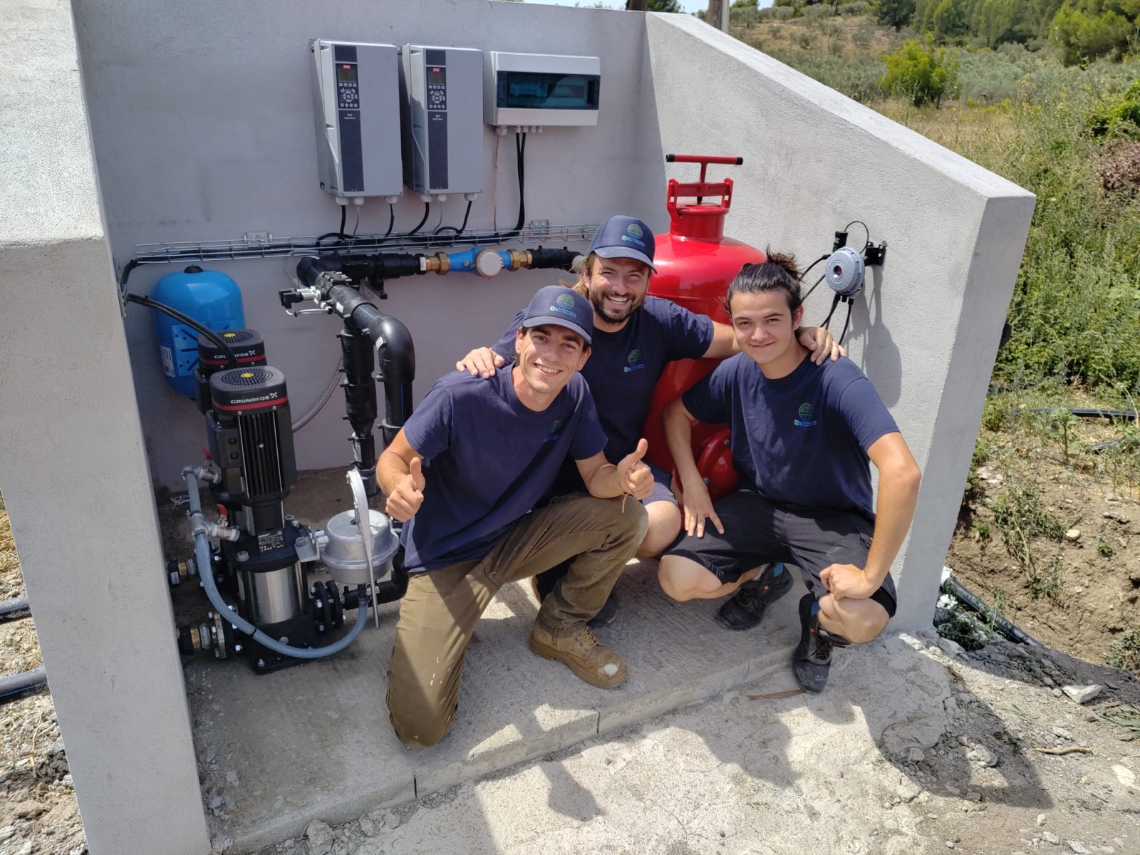 Trois membres de l'équipe devant un local de pompage (pompe, ballon, armoires de commande) — 2A AUTOMATISMES